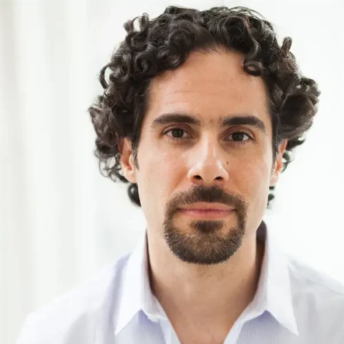 A man with curly dark hair, a goatee, and mustache, wearing a light-colored button-up shirt, looks directly at the camera against a bright, neutral background.