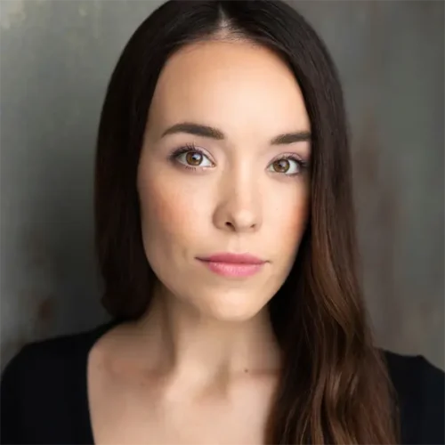A woman with long brown hair and light skin looks directly at the camera with a neutral expression, wearing a black top against a gray background.