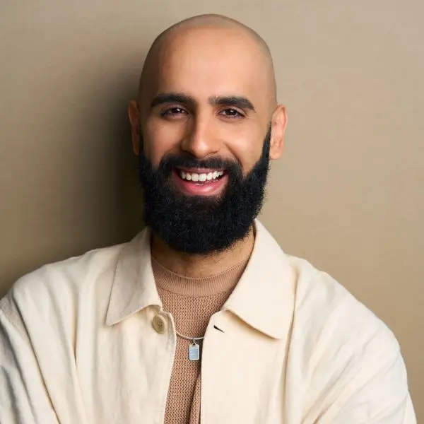 A bald man with a full beard and mustache smiles, wearing a beige jacket over a tan shirt, standing against a light brown background.