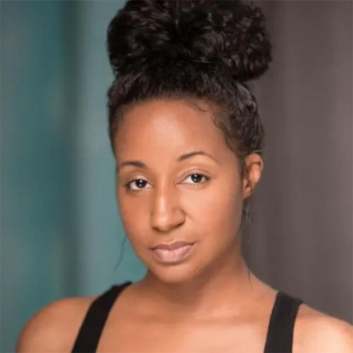 A woman with dark curly hair styled in a high bun, wearing a black tank top, looks at the camera against a blurred background.
