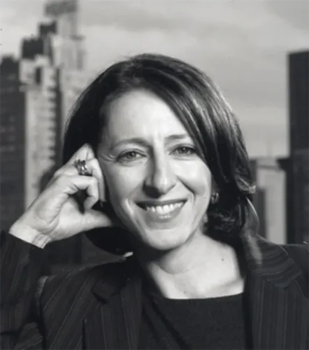 A woman with shoulder-length hair smiles while resting her head on her hand; she wears a striped blazer and sits in front of a cityscape background.