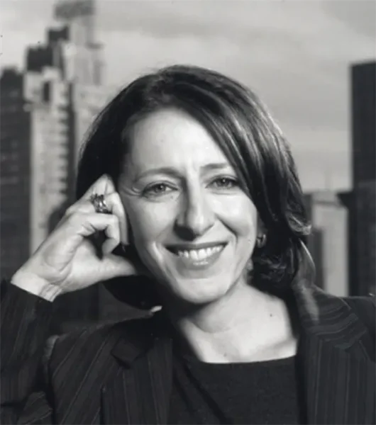 A woman with shoulder-length hair smiles while resting her head on her hand; she wears a striped blazer and sits in front of a cityscape background.