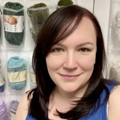 A woman with straight brown hair smiles in front of shelves displaying various colors of yarn.