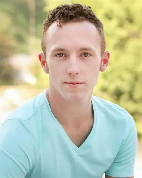 A young man with short curly hair wearing a light blue V-neck T-shirt is looking directly at the camera. The background is blurred greenery.
