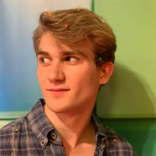 A young man with light brown hair and a plaid shirt looks to the side, with small pieces of confetti in his hair and on his shirt, standing against a colorful background.