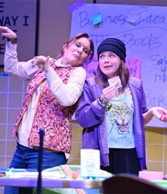 Two women stand on stage in front of a sign and a handwritten poster, one gesturing dramatically while the other holds a card and looks to the side.