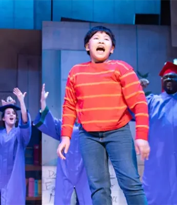 A child in a red striped shirt sings on stage, with two adults in purple graduation gowns and caps raising their arms in the background.