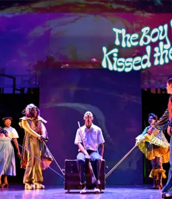 Five actors perform on stage with colorful costumes and props; a projected sign reads "The Boy Who Kissed the Sky" above them.