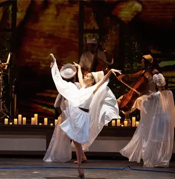 Dancers in flowing white costumes perform on stage, with musicians and lit candles in the background.