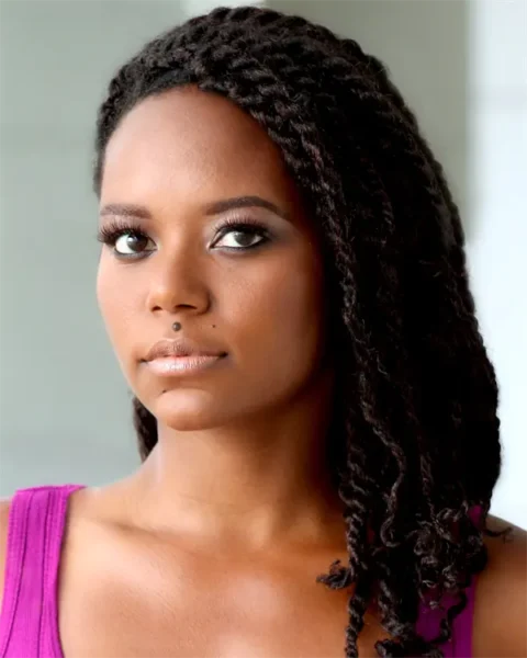 A woman with long, twisted dark hair and brown eyes, wearing a purple sleeveless top, looks at the camera against a neutral background.