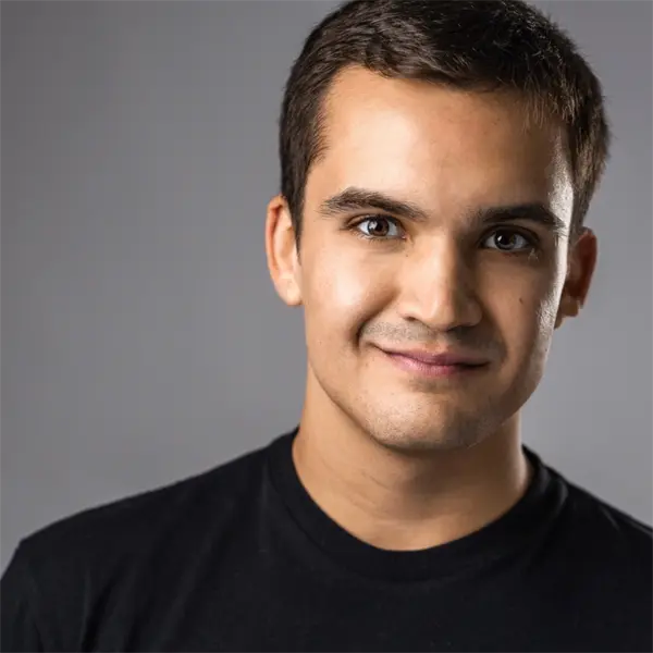 A young man with short dark hair, wearing a black t-shirt, stands in front of a plain gray background and smiles slightly at the camera.
