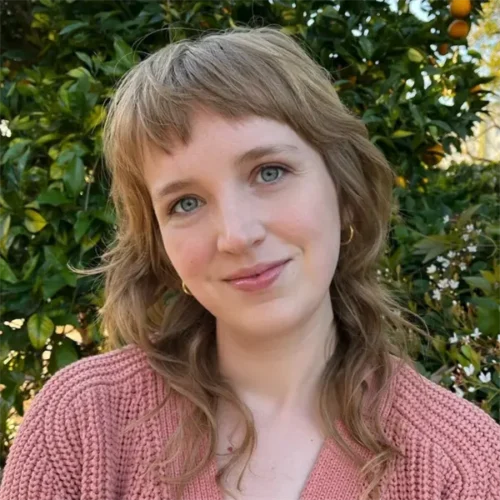 A person with wavy, light brown hair and blue eyes smiles softly, wearing a pink knit sweater and gold hoop earrings, standing outside in front of green foliage with small orange fruits.