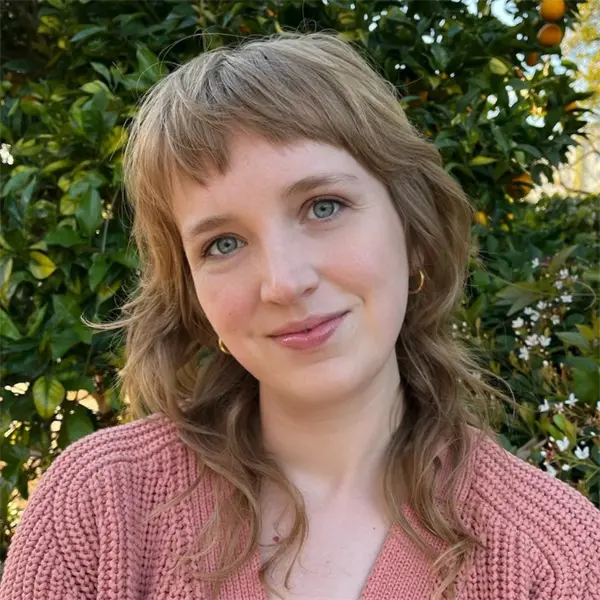 A person with wavy, light brown hair and blue eyes smiles softly, wearing a pink knit sweater and gold hoop earrings, standing outside in front of green foliage with small orange fruits.