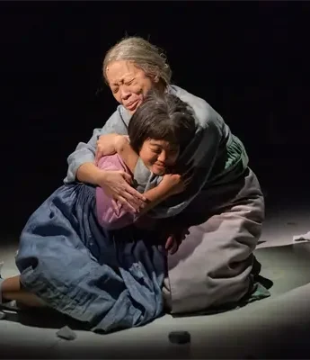 Two women sit on the floor under a spotlight, embracing each other tightly with emotional expressions; crumpled papers are scattered around them.