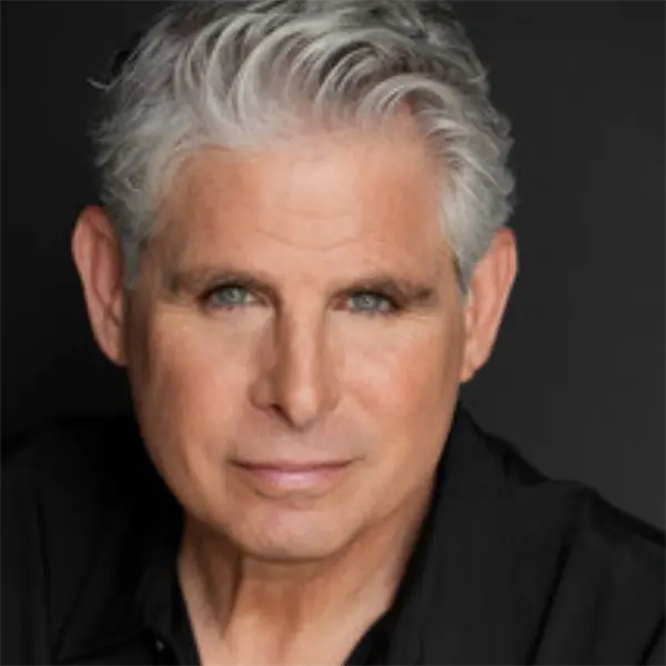 A middle-aged man with short gray hair and light eyes looks directly at the camera against a dark background. He is wearing a black shirt.