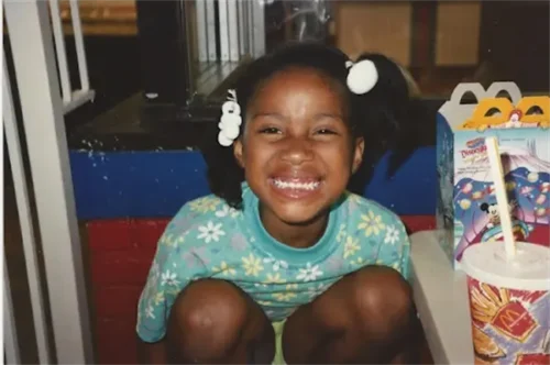 A young girl with pigtails sits and smiles widely at the camera next to a Happy Meal box and a McDonald's cup.