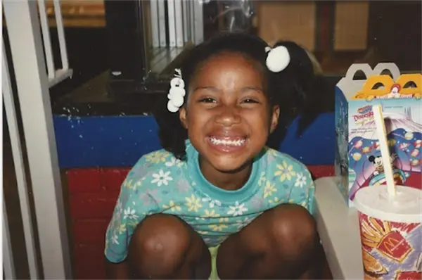 A young girl with pigtails sits and smiles widely at the camera next to a Happy Meal box and a McDonald's cup.