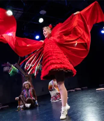 Performer in a bright red costume dances on stage with arms raised, while other performers in colorful outfits sit and watch in the background.