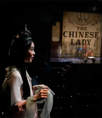 A woman in traditional Chinese attire stands in a dimly lit theater, looking toward a stage with a backdrop reading "The Chinese Lady.