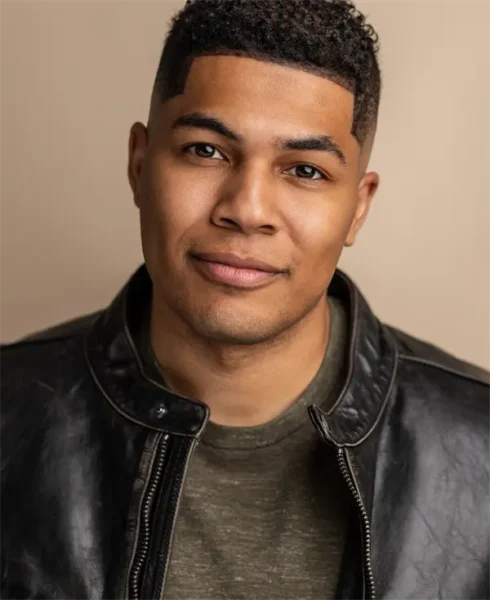 A man with short curly hair wearing a dark leather jacket and an olive green shirt, facing the camera with a neutral expression.