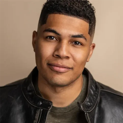 A man with short curly hair wearing a dark leather jacket and an olive green shirt, facing the camera with a neutral expression.