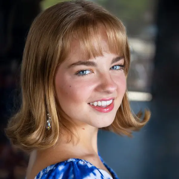 A young woman with shoulder-length blonde hair and blue eyes smiles at the camera, wearing a blue patterned top and dangling earrings.