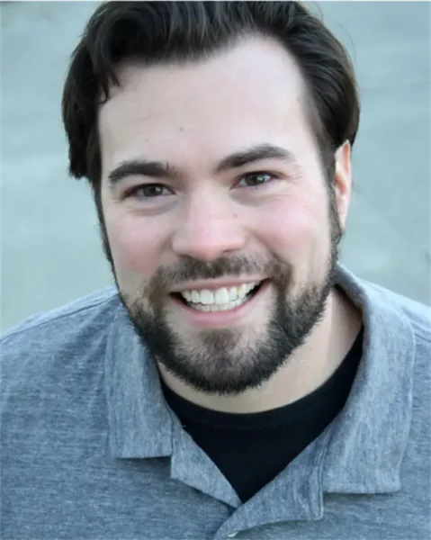 A man with dark hair and a beard is smiling at the camera, wearing a gray collared shirt over a black t-shirt. The background is out of focus.