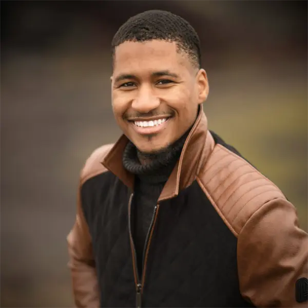A man wearing a black and brown jacket smiles at the camera with an outdoor blurred background.