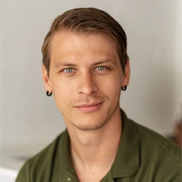 A man with short light brown hair, light stubble, and earrings, wearing a green collared shirt, looks directly at the camera against a neutral background.