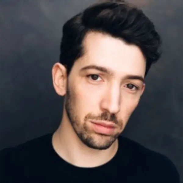 A man with short dark hair and facial hair is wearing a black shirt and looking at the camera against a dark background.