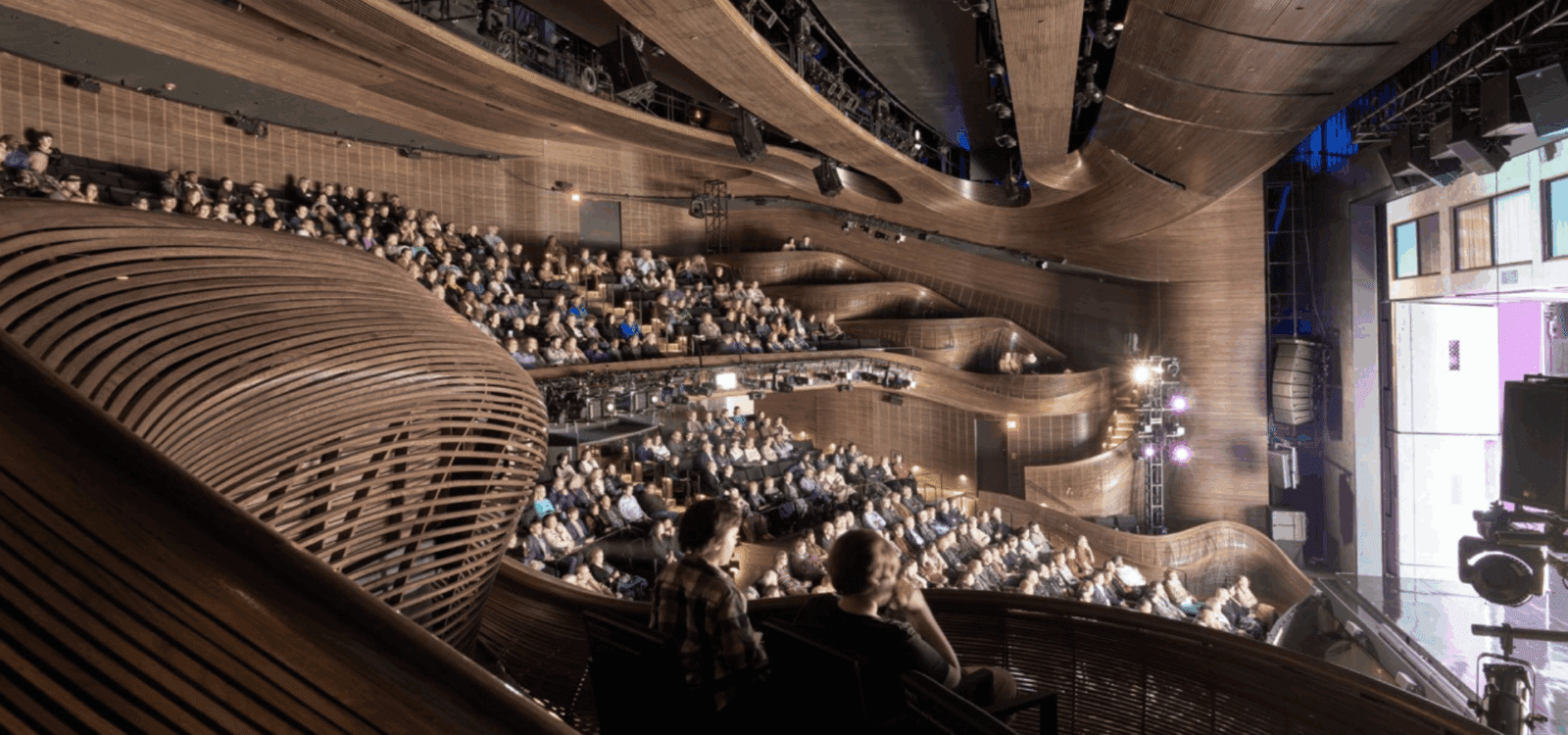 Audience seated in a modern, wood-paneled theater with multiple balcony levels, viewing a brightly lit stage on the right side of the image.