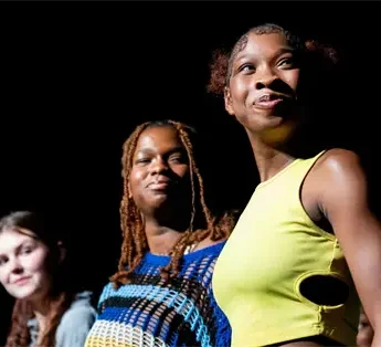 Five young women stand together onstage against a dark background, facing forward. One woman in a yellow top is in the foreground, slightly smiling.