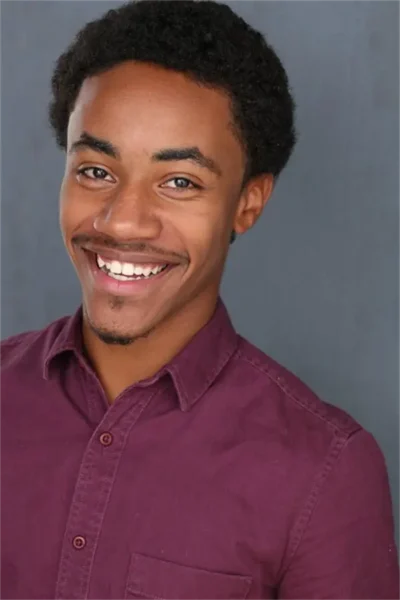 A young man with short curly hair smiles at the camera, wearing a maroon button-up shirt against a plain gray background.