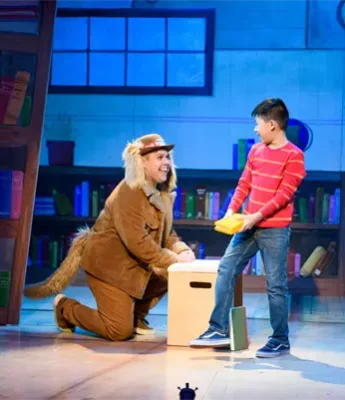 An adult in a lion costume kneels beside a young boy holding a yellow object on stage in a library-themed set.