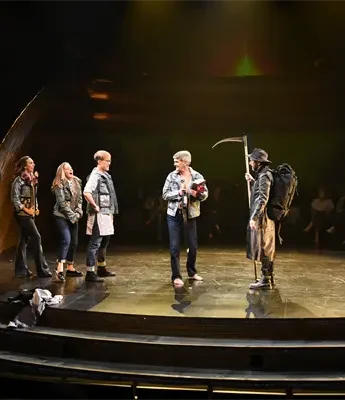 Actors stand on a circular stage under dramatic lighting, dressed in modern costumes, with one holding a scythe and others focused on him.