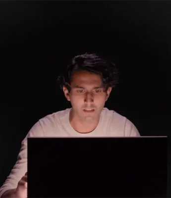 A person in a white shirt sits in front of a laptop, looking at the screen with a focused expression against a dark background.