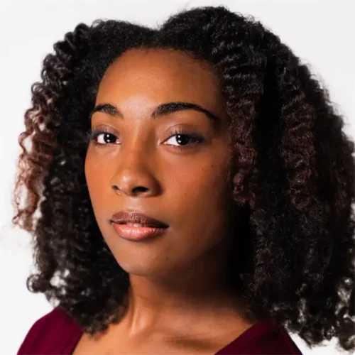 A woman with medium-dark skin and natural curly hair looks at the camera with a neutral expression, wearing a burgundy top against a plain white background.