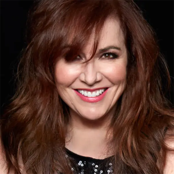 A woman with long, wavy auburn hair smiles at the camera against a black background, wearing a sleeveless black top with sequin details.