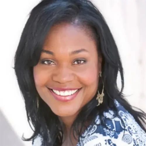 A woman with medium-dark skin and straight black hair smiles at the camera while wearing a patterned top and dangling earrings.