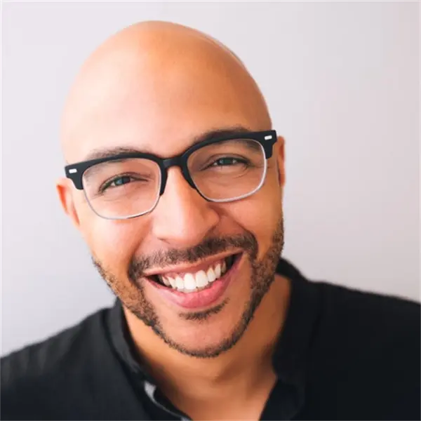 A smiling bald man with glasses and a trimmed beard wearing a black shirt, facing the camera against a plain light background.