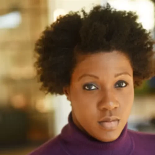 A woman with short curly hair wearing a purple turtleneck sweater looks directly at the camera with a neutral expression.