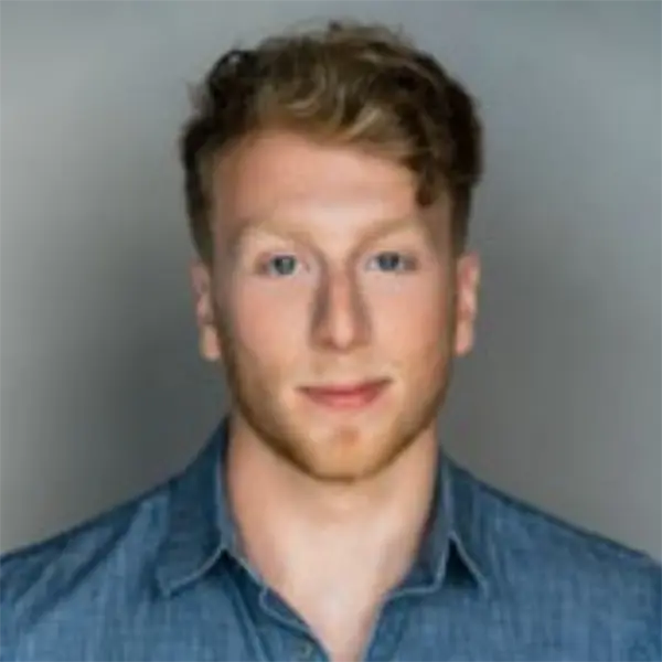 A man with short, wavy light brown hair and a beard is wearing a blue collared shirt and facing the camera against a plain background.