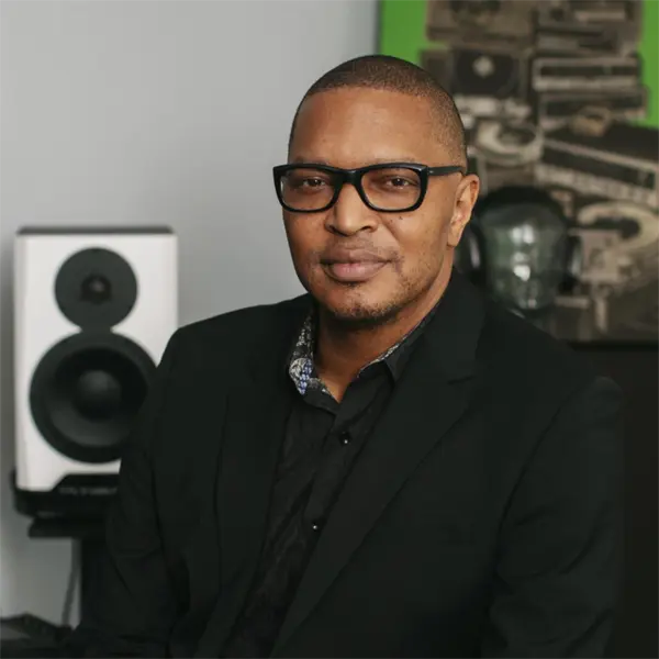 A man wearing glasses and a black suit sits indoors, with a speaker and artwork visible in the background.
