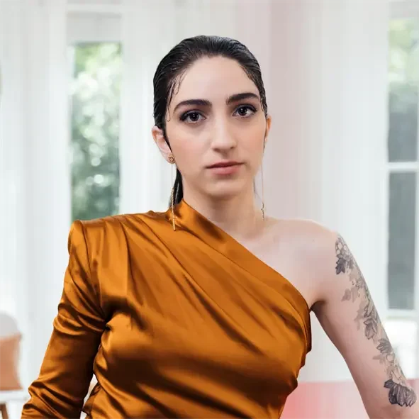 A person with dark hair in a slicked-back style wears a gold one-shoulder top, displaying a floral tattoo on their left arm, seated indoors with large windows in the background.