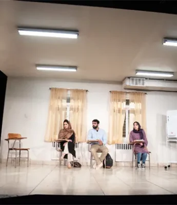 Five people are in a brightly lit classroom; four are seated facing a standing woman who appears to be speaking or presenting.