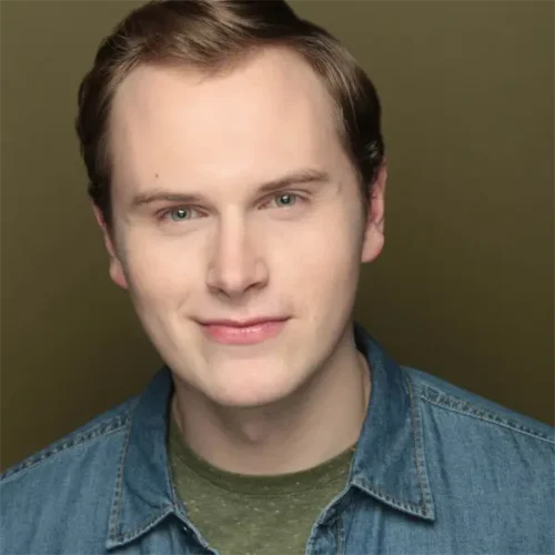 A young man with short light brown hair, fair skin, and blue eyes is wearing a denim shirt over a green t-shirt, smiling slightly against a plain, olive-brown background.
