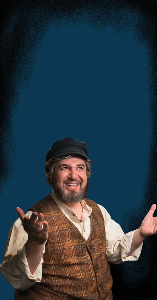 A man with a beard wearing a dark cap, white shirt, and brown plaid vest gestures with open hands and smiles against a blue background.