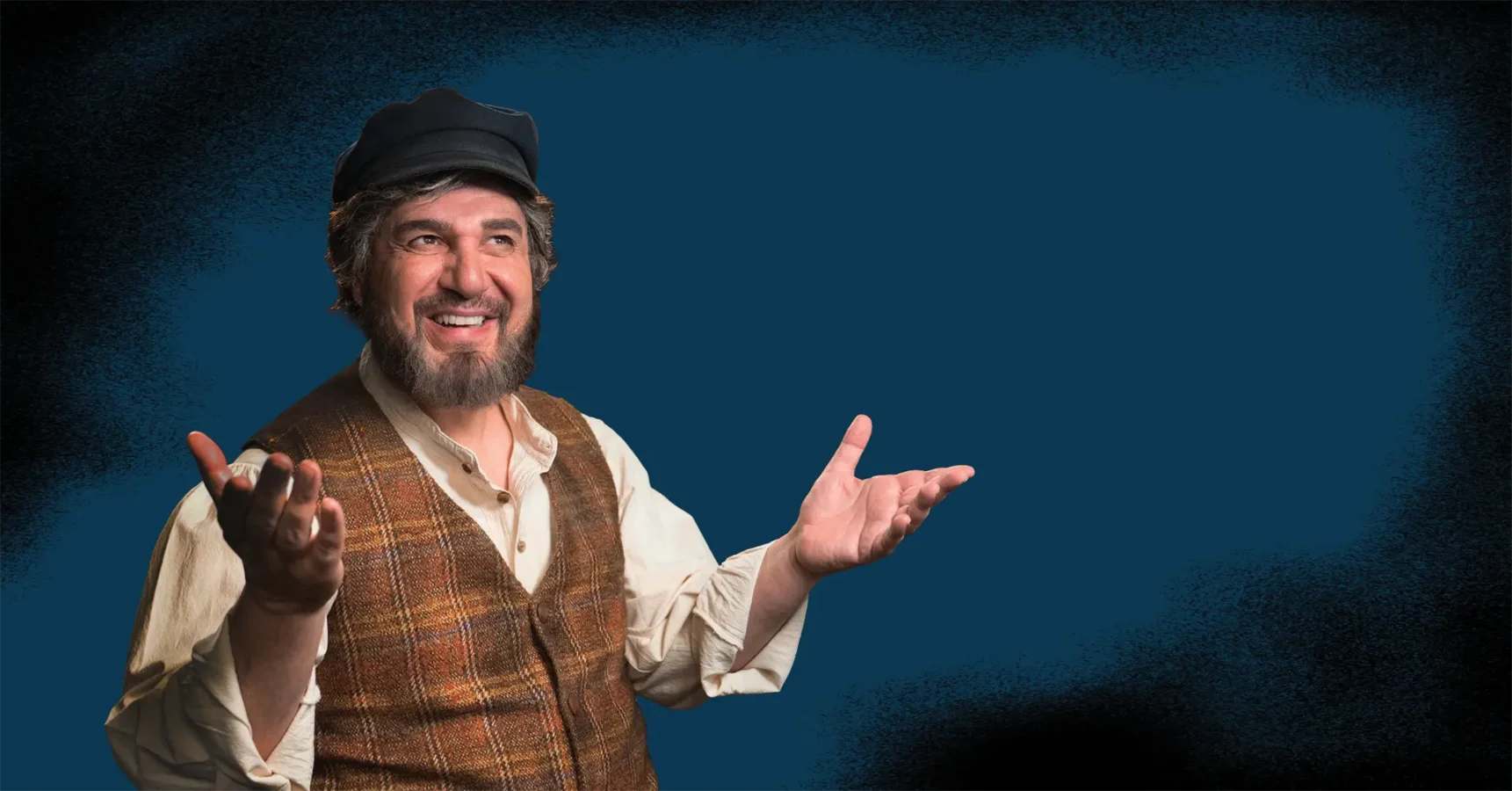 Man with a beard and hat, wearing a plaid vest over a shirt, smiles and gestures with both hands against a plain dark blue background.