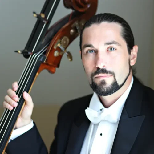 A man in formal attire with a white bow tie holds an upright bass, looking directly at the camera.