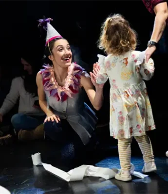 A woman in a party hat kneels and smiles at a young child in a floral dress, with another adult holding the child's hand on a dark stage.